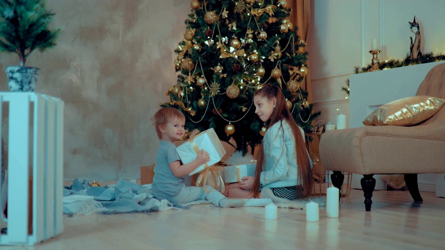 孩子们坐在圣诞树下观看礼物。男孩和女孩为惊喜而高兴，并试图打开礼物。圣诞树立在花环和玩具上。视频素材