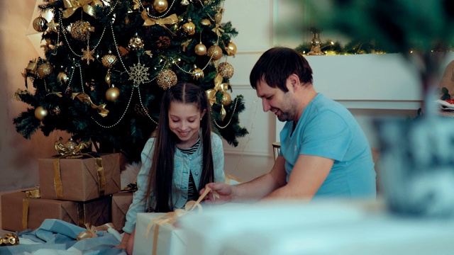 女孩和爸爸坐在新年树上，打开礼物的盒子。漂亮的新年房间。视频素材