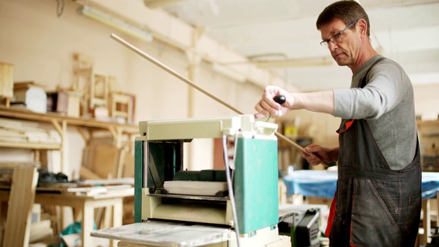 成熟的家具制造者在他的工作室里用木工机器打磨木板，眼镜和工装裤视频素材