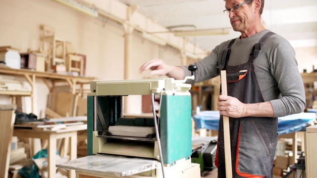 专业木匠在眼镜和工装裤使用木工机器打磨长木板在他的家具制造车间视频素材