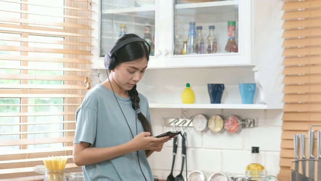 年轻美丽的亚洲女人戴着耳机和使用智能手机，而站在厨房视频素材