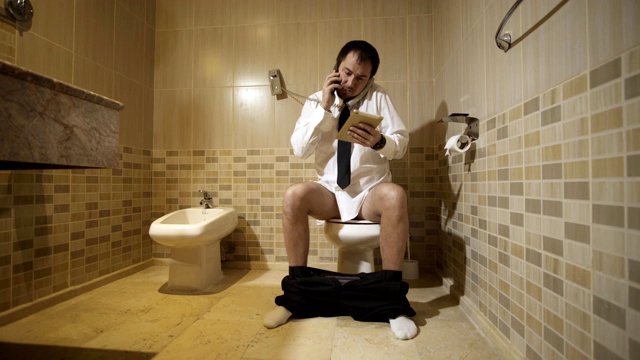 一个男人坐在豪华厕所的马桶上，一边用手机一边工作。FullHD视频下载