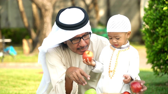 快乐的穆斯林小男孩和爷爷穿着传统服装在公园吃苹果早上视频下载