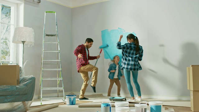 与小女儿共度家庭时光。年轻的爸爸和妈妈正在用油漆滚筒跳舞和胡闹。墙漆颜色为浅蓝色。家里的房间准备装修。视频下载