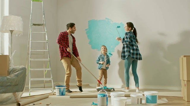 与小女儿共度家庭时光。年轻的爸爸和妈妈正在用油漆滚筒跳舞和胡闹。墙漆颜色为浅蓝色。家里的房间准备装修。视频下载