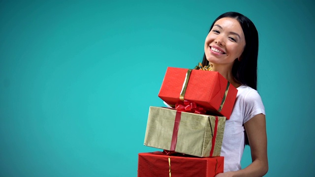 微笑的女孩拿着许多礼盒，节日的礼物，站在蓝色的背景视频购买