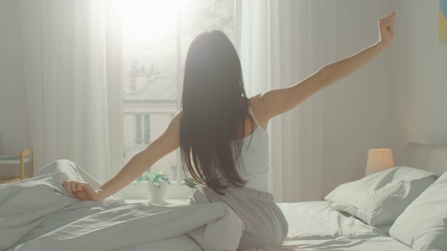 美丽的黑发女人早上慢慢醒来，伸展身体，从床上爬起来，阳光从大窗户照在她身上。快乐的年轻女孩迎接新的一天。视频下载