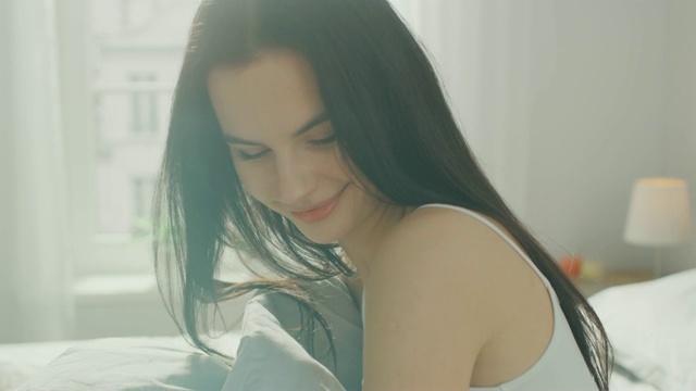 美丽的黑发女人在早晨慢慢醒来，坐在床上，迷人的微笑。阳光从窗口照射在一个快乐的醒来的年轻女孩。视频素材
