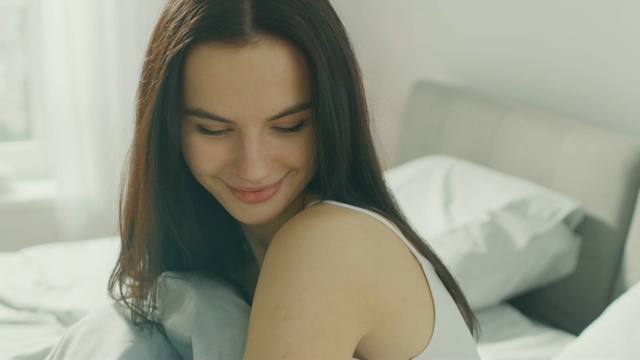 性感的年轻女孩在白色坐在床上，迷人的微笑。美丽的黑发女人躺在床上，阳光照耀着她。视频素材
