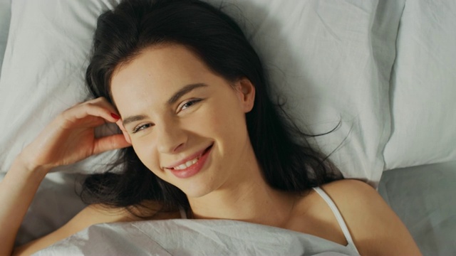 美丽的年轻的黑发女人迷人的画像在她的床上醒来，性感的微笑，她的脸被温暖的早晨的太阳照亮。甜蜜和温暖。自上而下旋转相机拍摄。视频素材