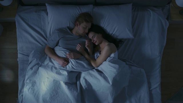 幸福的年轻夫妇拥抱在床上睡觉在晚上。美女和帅哥睡在一起，甜蜜地拥抱在一起。自顶向下。视频下载