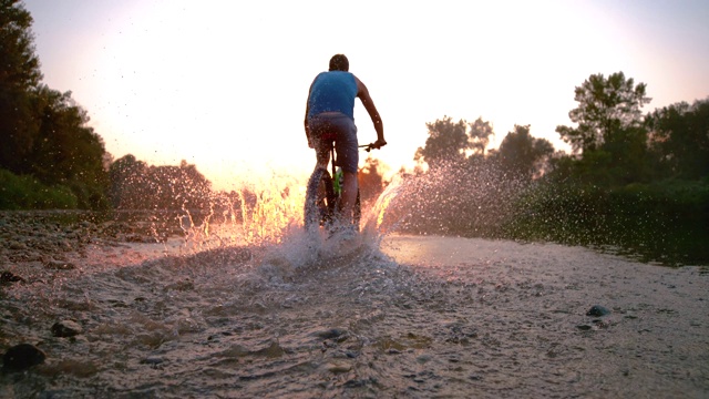 慢镜头:一名运动员骑着自行车在浅溪中向夕阳走去。视频素材