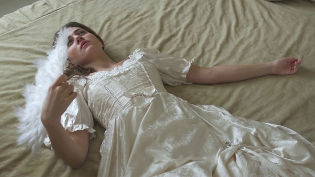 年轻漂亮的女人，穿着白色的老式舞会礼服，躺在巨大的金色床上，挥动着羽毛扇。视频下载