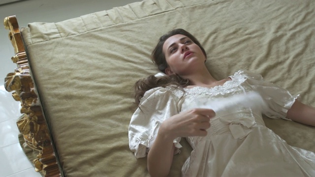 穿着舞会礼服的年轻疲惫的女人躺在床上，挥舞着一把扇子。视频下载