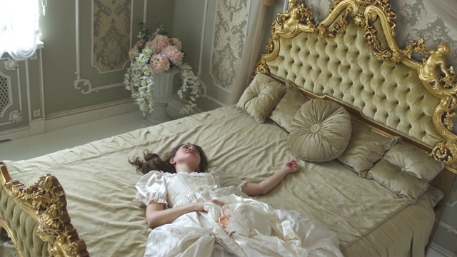 年轻女子在舞会礼服落在金色装饰的床上拿着丝绸帽子。女孩又累又困视频下载