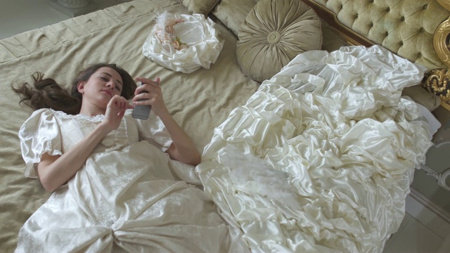 年轻疲惫的女人在舞会礼服躺在金色装饰的床上，用手机短信近距离。视频下载