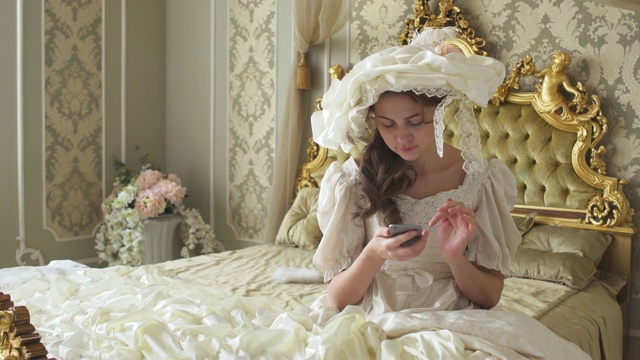 可爱的年轻女子穿着舞会礼服坐在金色装饰的床上，用手机发短信视频下载