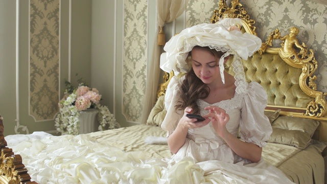 年轻女子穿着舞会礼服坐在金色装饰的床上，在手机上发短信。公主使用小工具视频下载