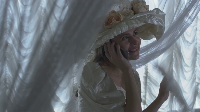 一个年轻漂亮的女人，穿着白色的复古舞会裙，戴着白色的帽子，站在透明窗帘的背景上，挥舞着扇子，在讲手机视频素材