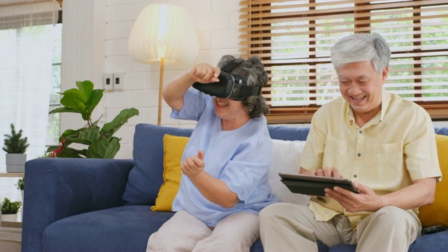 一对亚洲老年夫妇坐在家里的沙发上，戴着虚拟现实眼镜，拿着遥控器。高级快乐时刻视频素材