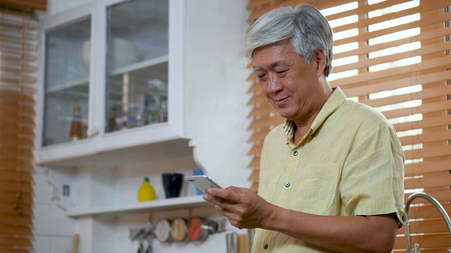 肖像高级亚洲男子使用智能手机在厨房快乐和微笑在家里。慢动作视频素材