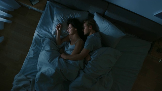 夜景床:迷人的年轻夫妇睡在一起，互相抱在怀里，拥抱。蓝色的夜间颜色和寒冷微弱的灯柱灯光透过窗户。视频下载