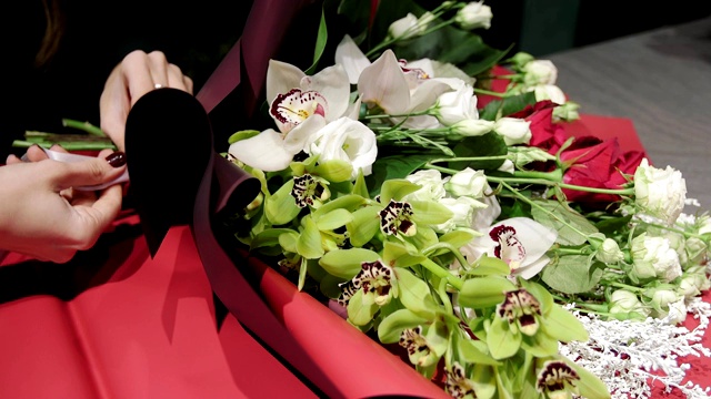 花商将丝带系在一束美丽的花从兰花，红和白玫瑰和一枝黄花在纸，手近距离。视频素材