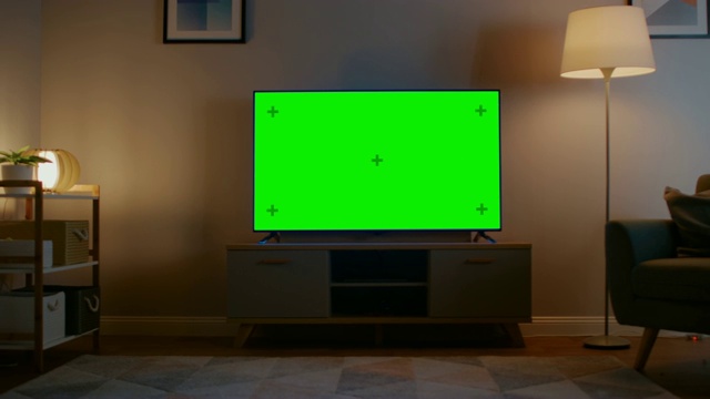 缩小镜头的电视水平绿色屏幕模拟起来。舒适的夜晚客厅，家里有一把椅子和打开的灯。视频素材