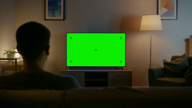 戴眼镜的年轻人正坐在沙发上看水平绿色屏幕模拟电视。现在是晚上，家里的房间有工作灯。视频素材