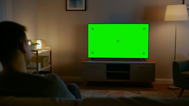 戴眼镜的年轻人正坐在沙发上看水平绿色屏幕模拟电视。现在是晚上，家里的房间有工作灯。视频素材