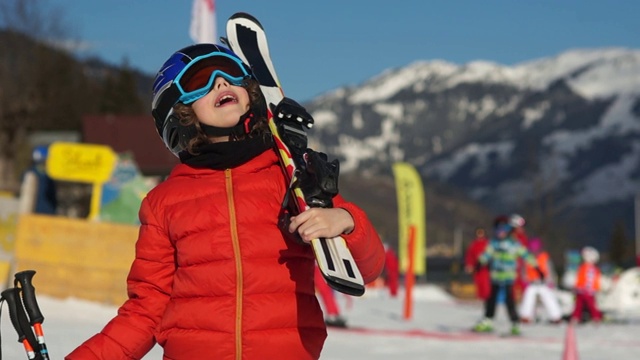 滑雪胜地的男孩。一个穿着全套滑雪装备的孩子，穿着红色夹克，戴着眼镜和头盔望着天空。阳光灿烂视频素材