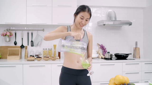 运动的亚洲女人在厨房喝苹果汁，美丽的女性在运动服装使用有机水果大量营养在家里自己做苹果汁。健康食品的概念。视频下载
