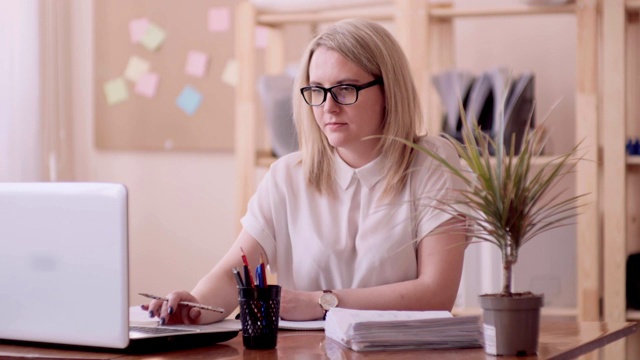 白种人外表的金发女孩戴着眼镜右手在办公室鲜花的右边坐在笔记本检查纸和用铅笔写笔记。视频素材