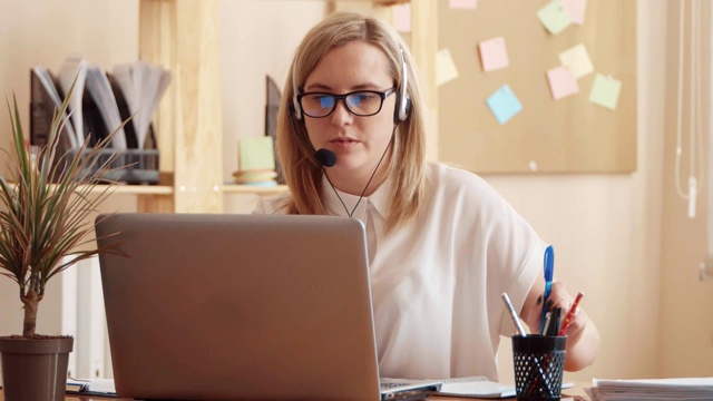 呼叫中心员工在办公室工作。一个金发姑娘戴着眼镜坐在办公室的桌子旁处理文件。她拿起电话，在键盘上打字视频素材