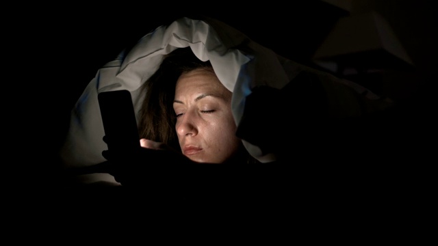 年轻女人在床上玩智能手机。一个女人在睡觉前用4k屏幕看手机视频下载