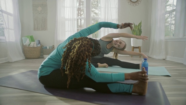 在家庭瑜伽室里，两个女人通过坐着的侧边伸展来模仿对方的动作。视频素材