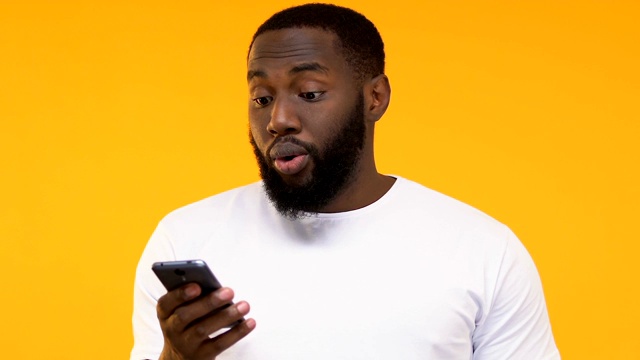 一名惊讶的黑人看着智能手机，手机应用更新，黄色背景视频素材