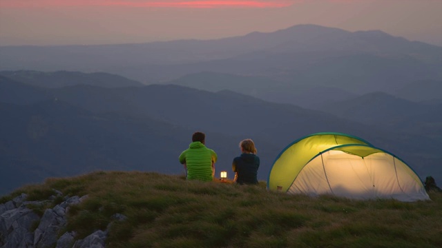 慢镜头:一对在阿尔卑斯山露营的游客夫妇在一个平静的夜晚坐在灯笼旁聊天视频素材