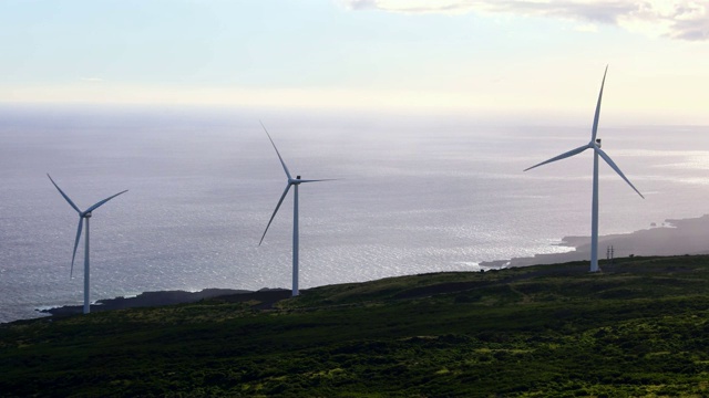 三个白色的风力涡轮机在一个绿色的山上俯瞰大海视频素材
