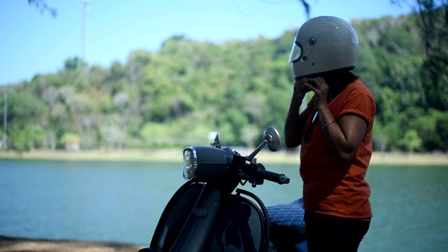 年轻的女性戴上头盔靠近她的摩托车，在湖边。视频下载