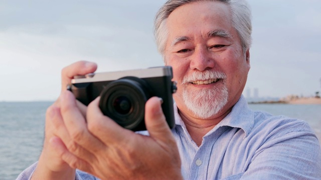 年长的亚洲男人用相机在海滩上与快乐的情绪。有生活方式、退休和科技观念的人。视频下载