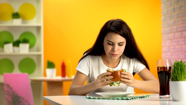 饥饿的年轻女士吃美味的芝士汉堡在咖啡馆，软饮料杯在桌子上视频素材