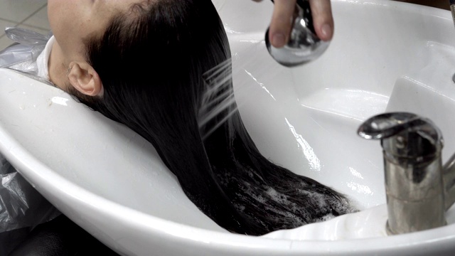在理发店里，美发师用从淋浴间流出的强劲水流在白色的水槽上给女孩洗头发。头发护理。关闭了。4 k。视频下载