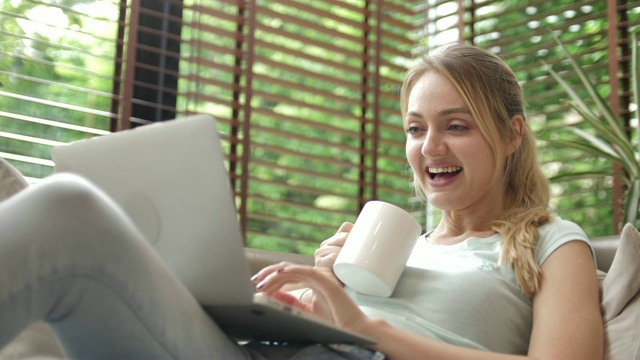 在客厅里用笔记本电脑的女人，微笑着拿着咖啡杯视频素材