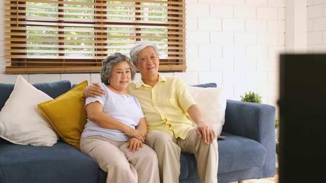 亚洲资深夫妇坐在沙发上，用遥控器在家里的客厅里换频道和看电视。从模糊前景电视平移视频下载