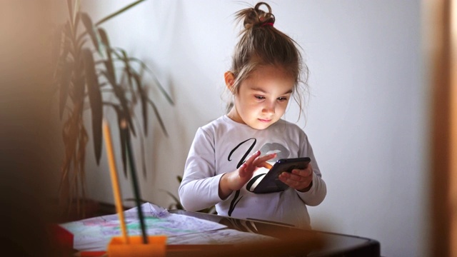 可爱的小女孩在她的房间，呆在桌子附近使用智能手机。-股票视频…视频素材