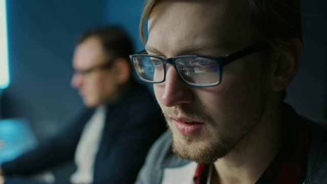 一个聪明专注的年轻人戴着眼镜在台式电脑上工作的肖像。背景模糊的办公室视频素材