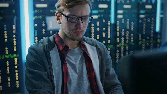 一个聪明专注的年轻人戴着眼镜在台式电脑上工作的肖像。在后台技术部办公室配备功能性数据服务器机架。视频素材