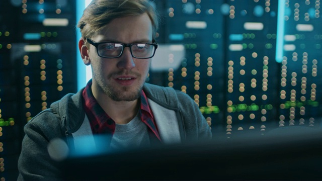 一个聪明专注的年轻人戴着眼镜在台式电脑上工作的肖像。在后台技术部办公室配备功能性数据服务器机架。视频素材