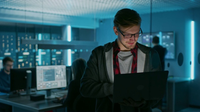 一个聪明的专注的年轻人的肖像戴着眼镜手持笔记本电脑。在后台技术部办公室，有专家工作和功能数据服务器机架视频素材